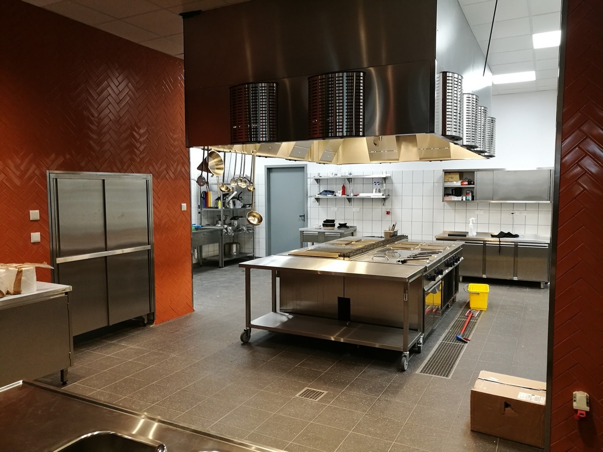 Profesjonalna kuchnia gastronomiczna ze stali nierdzewnej, widoczny duży okap, stół roboczy na kółkach, ściana wyłożona ceglastą glazurą w jodełkę, oraz sprzęt kuchenny.
