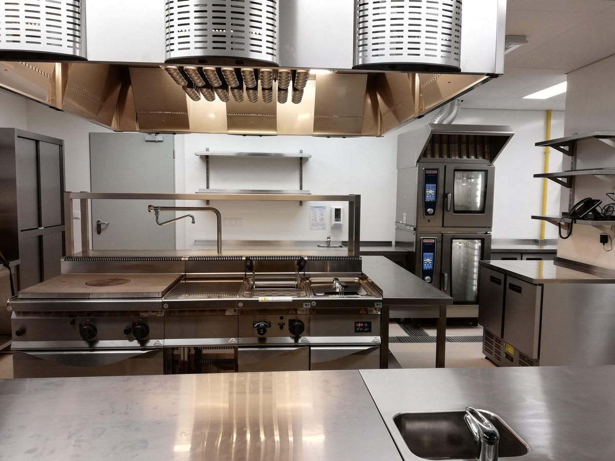 Profesjonalna kuchnia gastronomiczna ze stali nierdzewnej: piec konwekcyjny, płyta grzewcza z okrągłym palnikiem, frytownica i okap wentylacyjny z filtrami lamelkowymi, widok z perspektywy blatu...