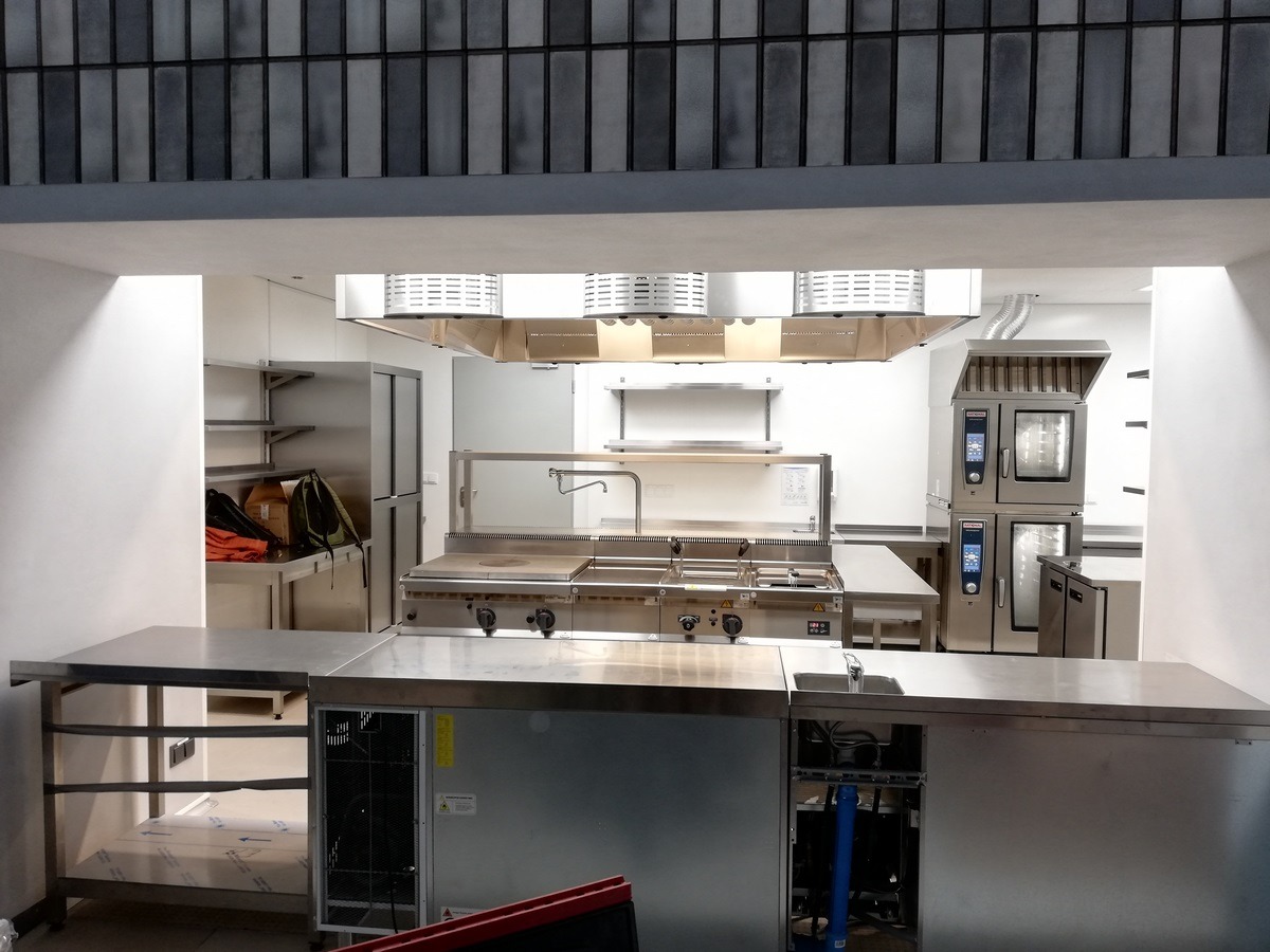 Profesjonalna kuchnia gastronomiczna ze stali nierdzewnej, widok przez okno, z widocznymi piecami konwekcyjnymi, płytą grzewczą, okapem i stołami roboczymi.