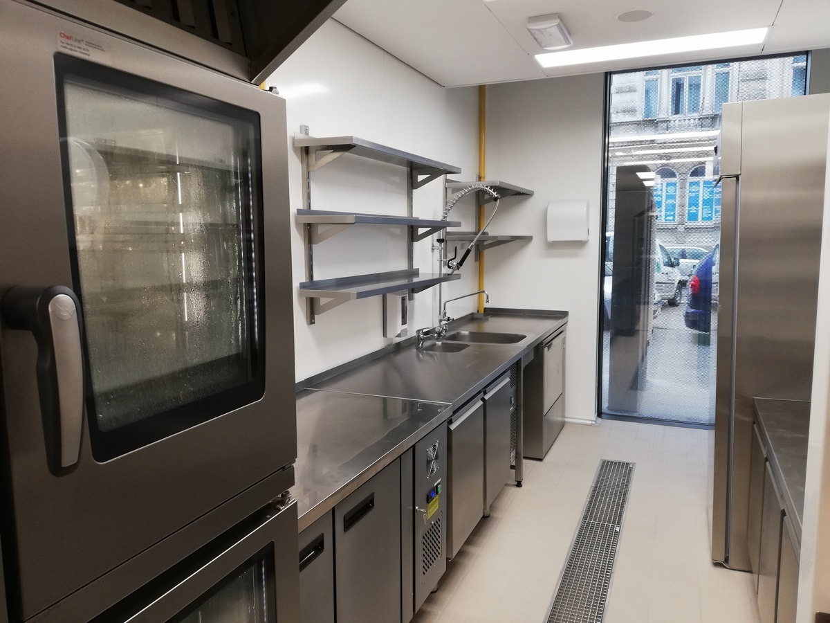 Profesjonalna kuchnia ze stali nierdzewnej wyposażona w piec konwekcyjny, zlewozmywak z baterią przemysłową, chłodziarki podblatowe i półki ścienne, widok z okna na budynek miejski.