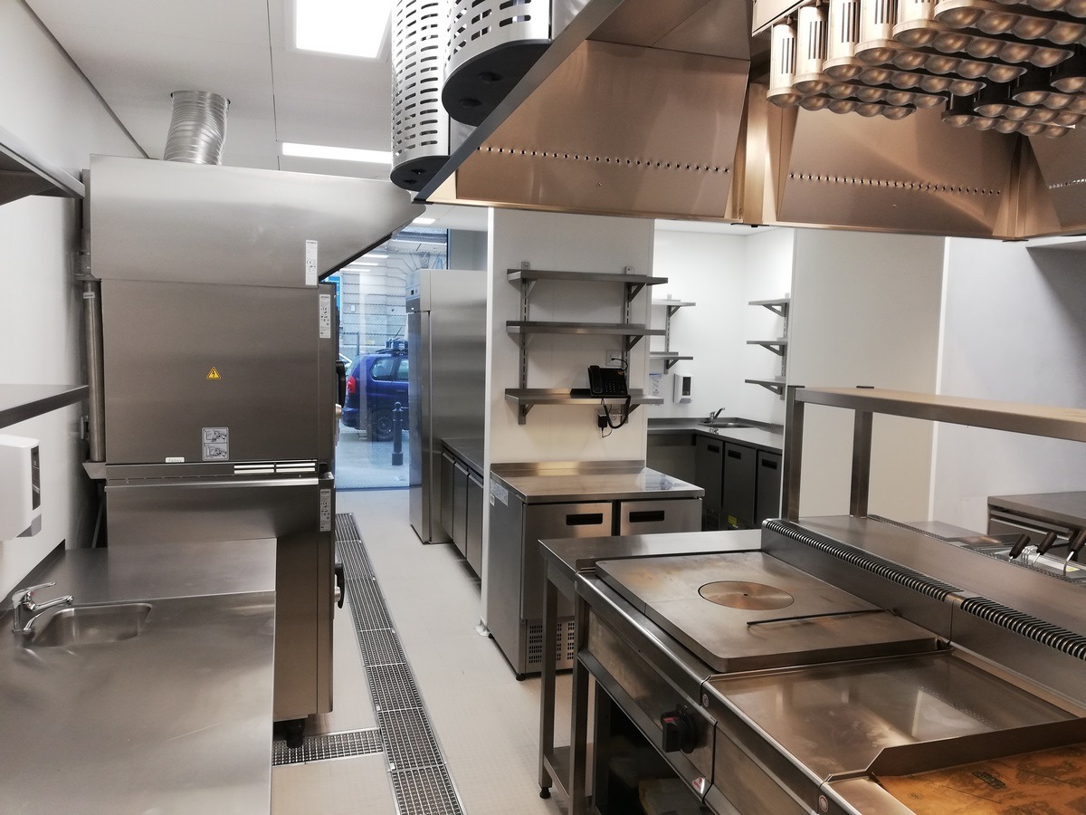Profesjonalna kuchnia gastronomiczna ze stali nierdzewnej, widoczne urządzenia do gotowania, okap z oświetleniem punktowym i system wentylacji, minimalistyczne półki na ścianie, podłoga z odpływem...