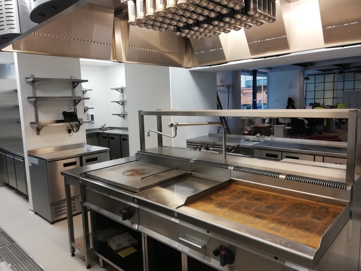 Profesjonalna kuchnia gastronomiczna ze stali nierdzewnej, widoczne płyty grzewcze, okap, szafki chłodnicze i półki wiszące, wnętrze lokalu w trakcie wykończenia.