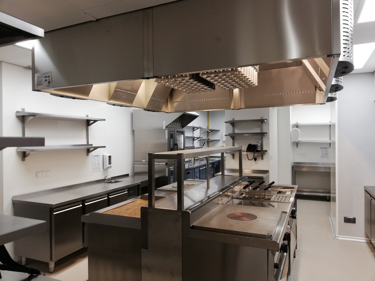 Profesjonalna kuchnia gastronomiczna ze stali nierdzewnej, widoczny duży okap nad stanowiskiem do gotowania, półki i zlewozmywak w tle.