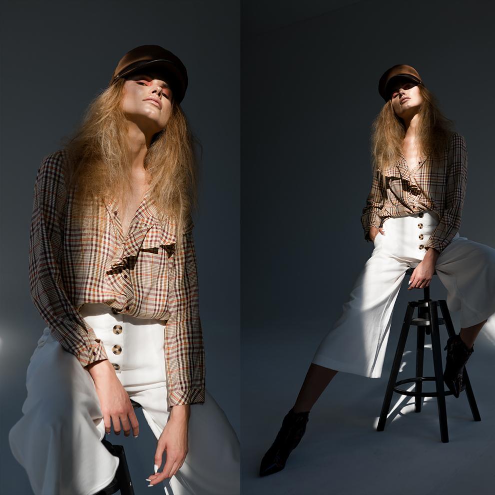 Portret kobiety siedzącej na stołku w studio, oświetlonej mocnym światłem, w kraciastej koszuli, białych spodniach z guzikami i brązowej czapce.