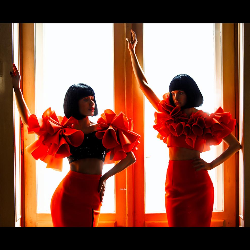 Dwie modelki w identycznych czerwonych strojach z dużymi falbanami pozują na tle jasnych drzwi, jedna z podniesioną ręką, druga z ręką na biodrze, w mocnym oświetleniu.