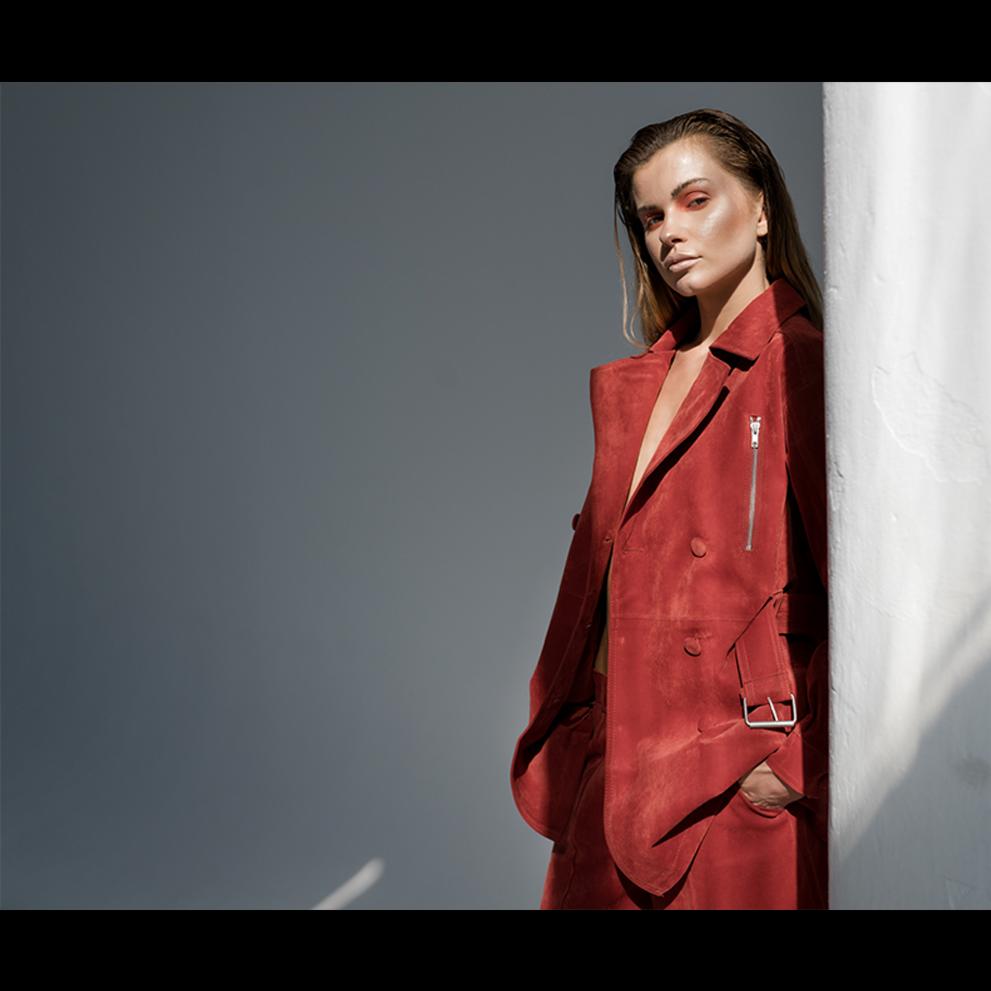 Kobieta w zamszowym, czerwonym garniturze pozuje oparta o białą kolumnę w ostrym świetle, z intensywnym makijażem oczu.