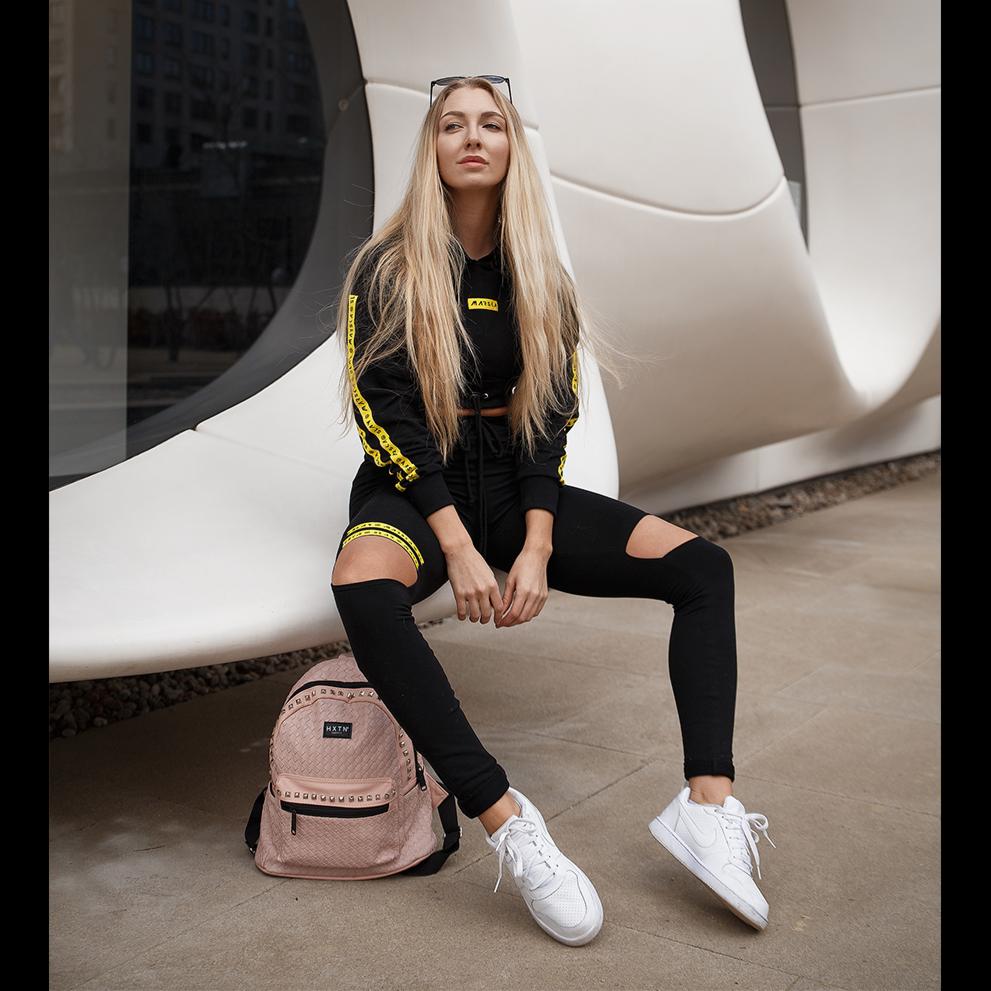 Kobieta z długimi blond włosami w czarnym dresie z żółtymi paskami, siedząca na białej, futurystycznej konstrukcji na tle budynku, z różowym plecakiem i białymi butami.