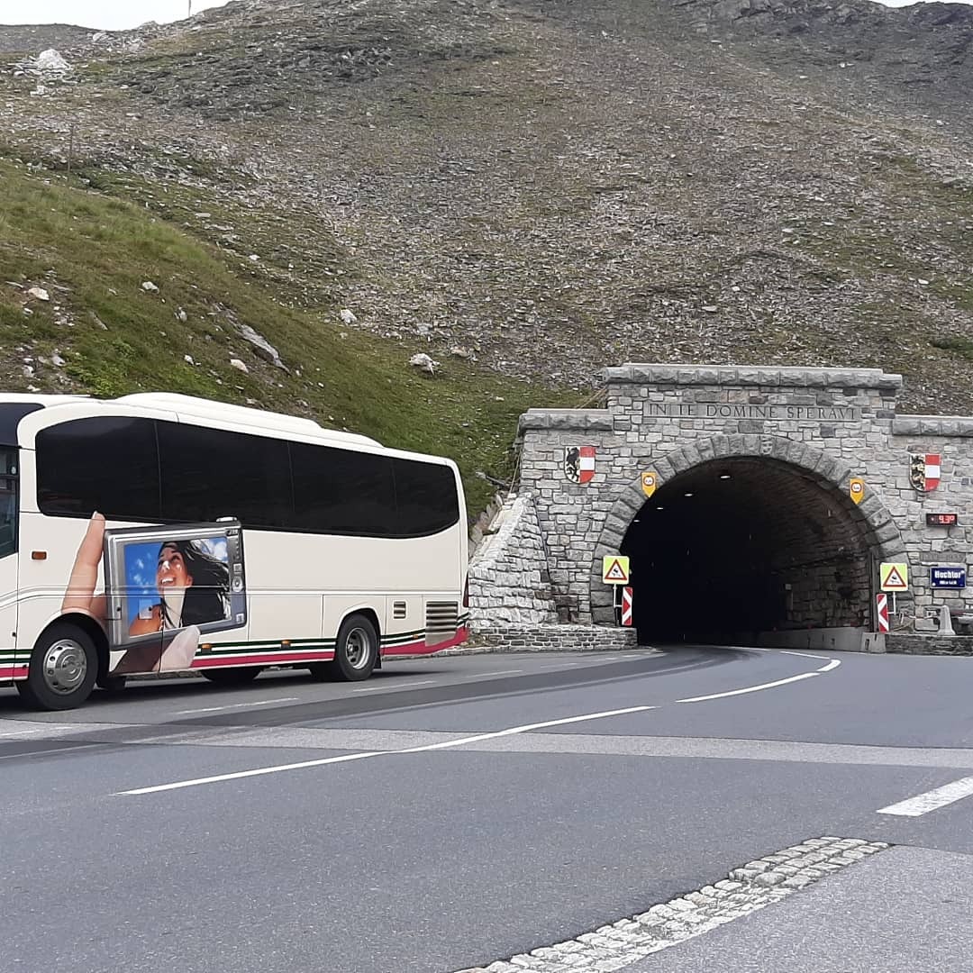 Autokar turystyczny z reklamą na boku, stojący przed kamiennym tunelem w górach.