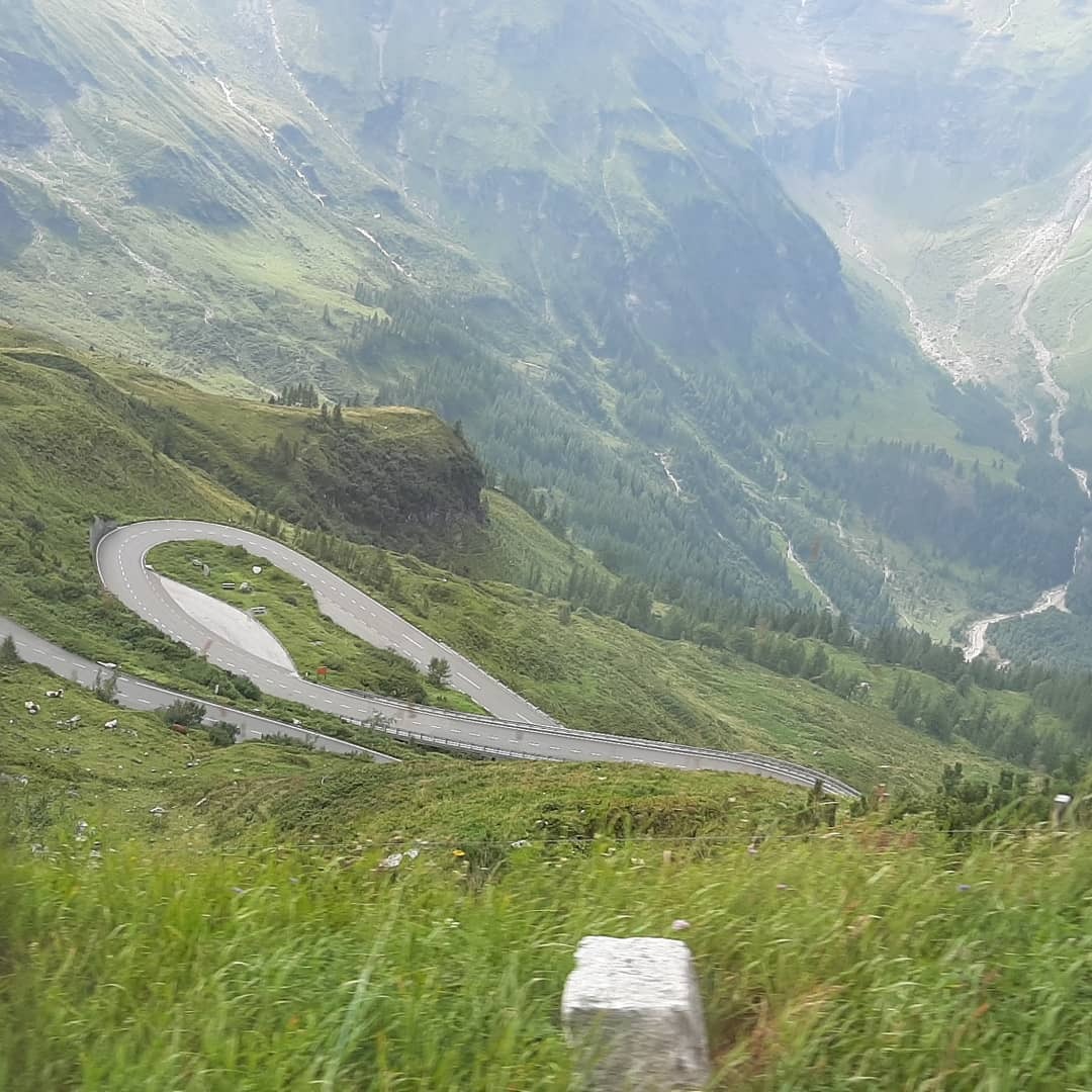 Widok z góry na serpentyny drogi w górach, otoczone zielenią i mgłą, z betonowym słupkiem w dolnym rogu kadru.