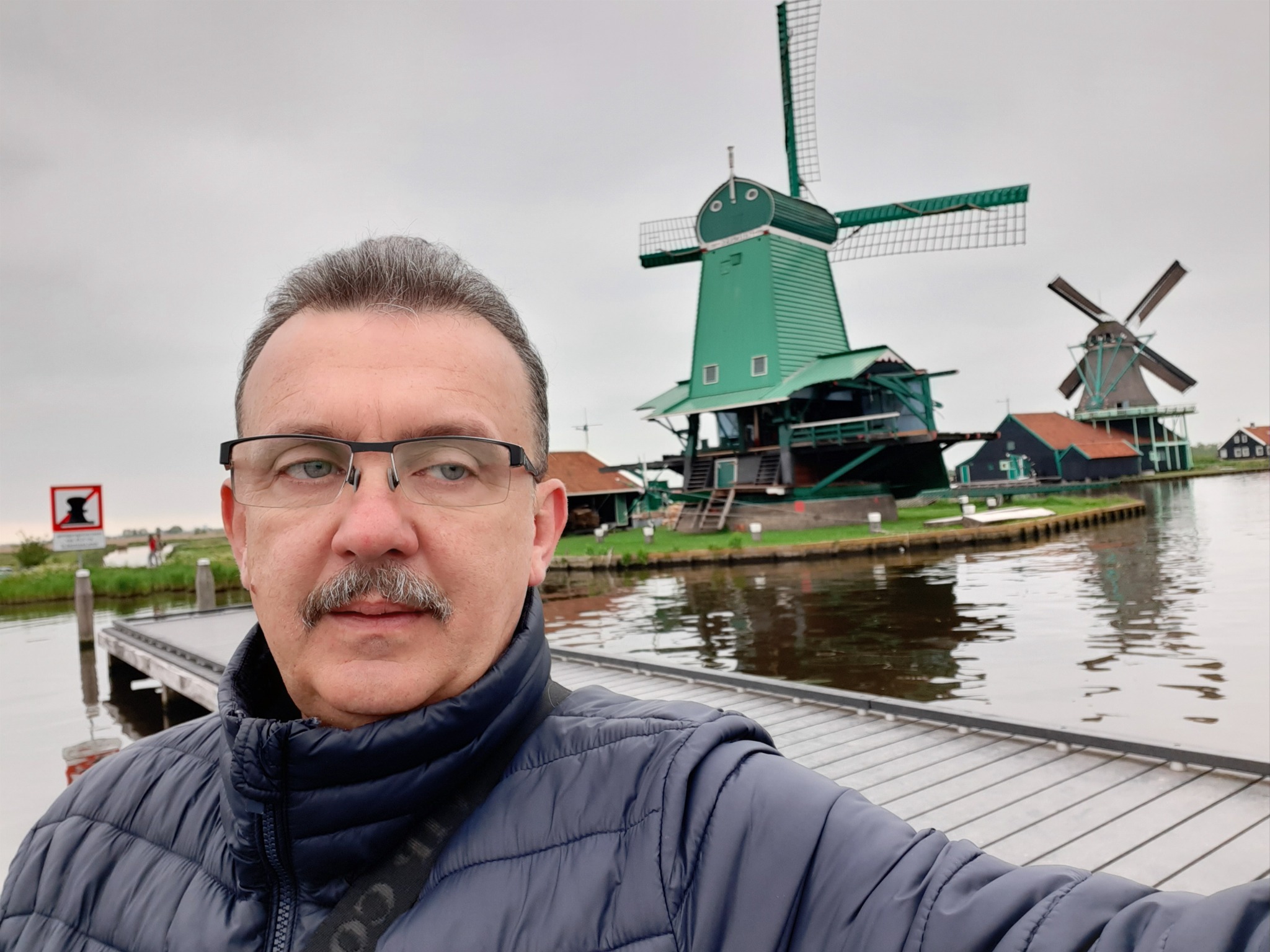 Mężczyzna w okularach i kurtce na drewnianym pomoście z wiatrakami w tle.