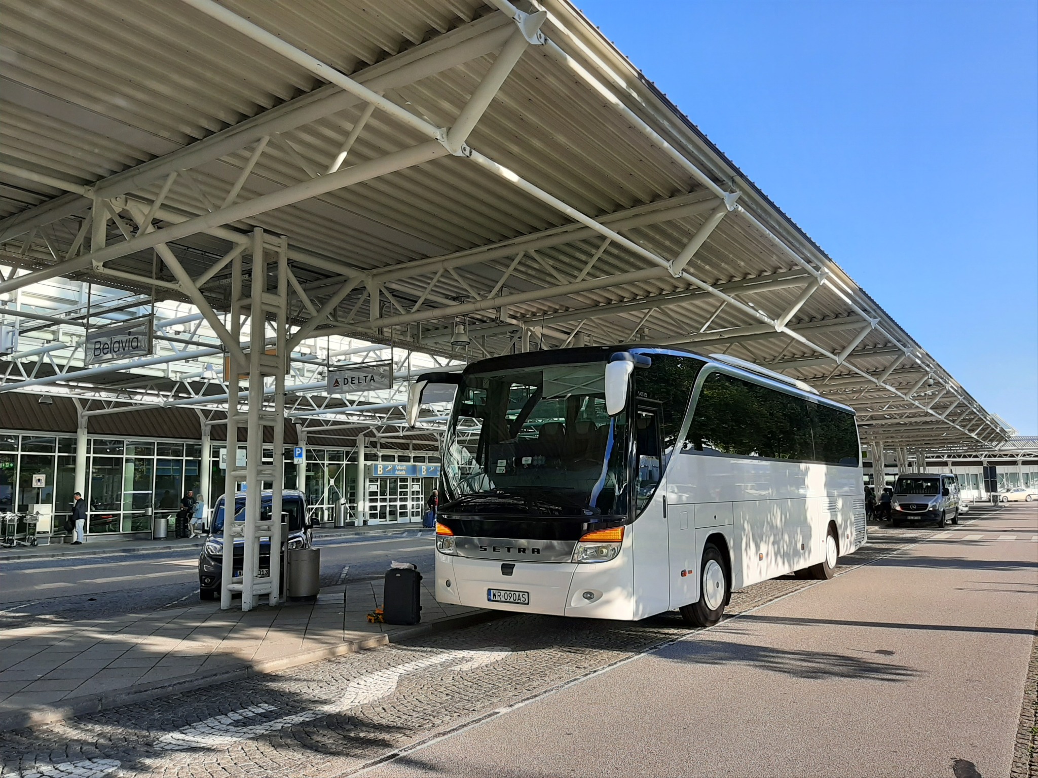 Biały autokar SETRA z otwartymi drzwiami, zaparkowany pod zadaszeniem przystanku autobusowego na lotnisku, obok bagaż i inne pojazdy.