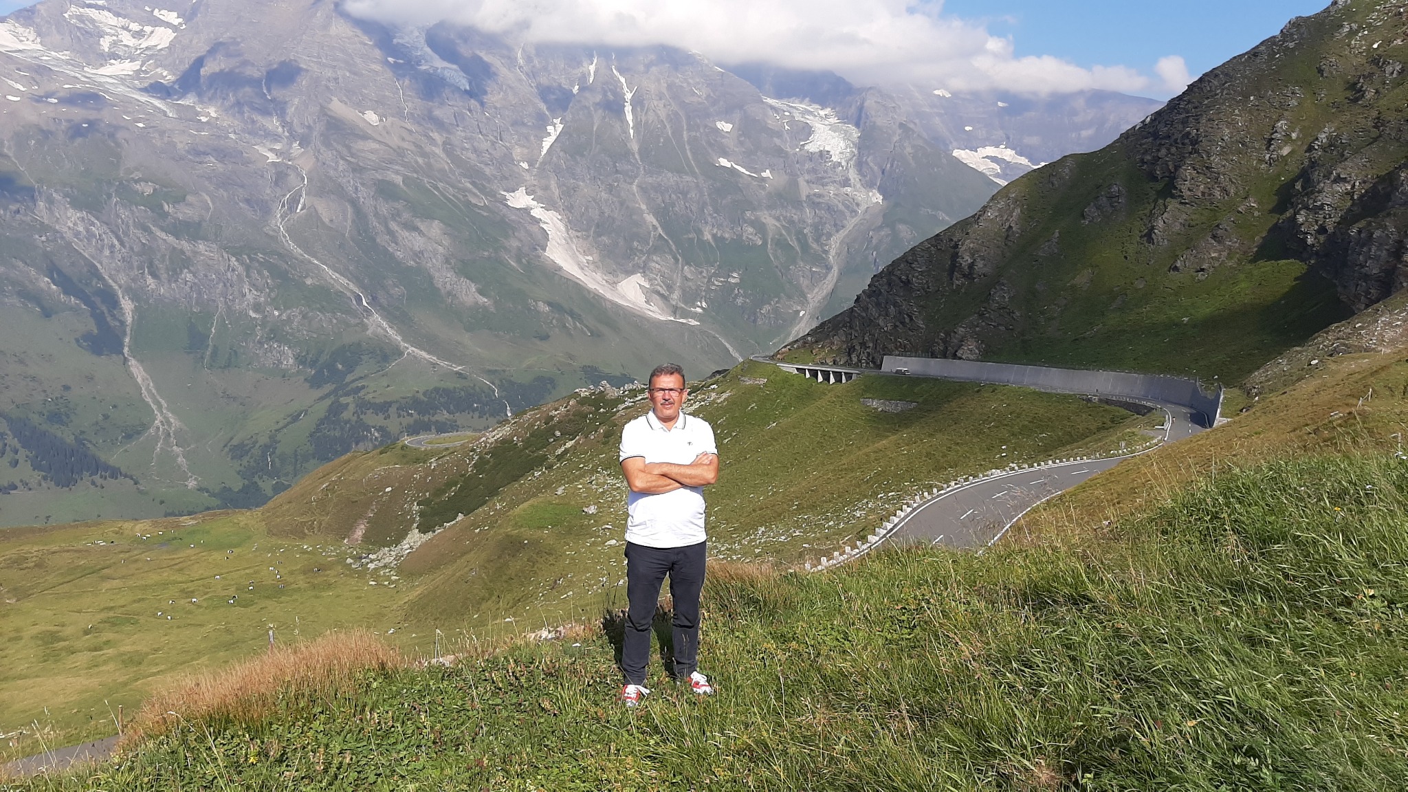 Mężczyzna w okularach i białej koszulce, stojący na tle górskiej drogi z tunelem i ośnieżonych szczytów