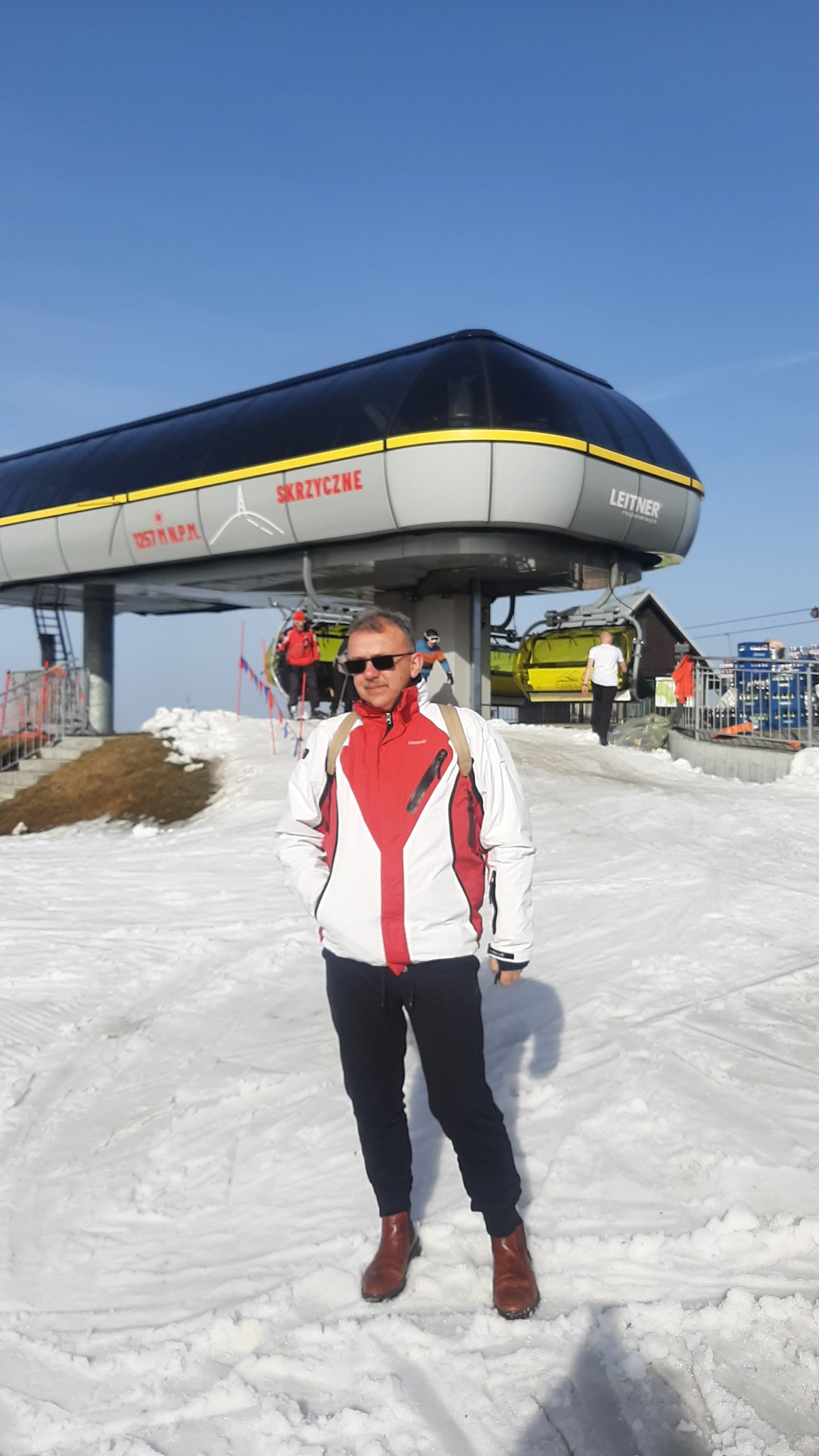 Mężczyzna w okularach przeciwsłonecznych, kurtce narciarskiej i brązowych butach stoi na śniegu na tle stacji kolejki linowej Skrzyczne.