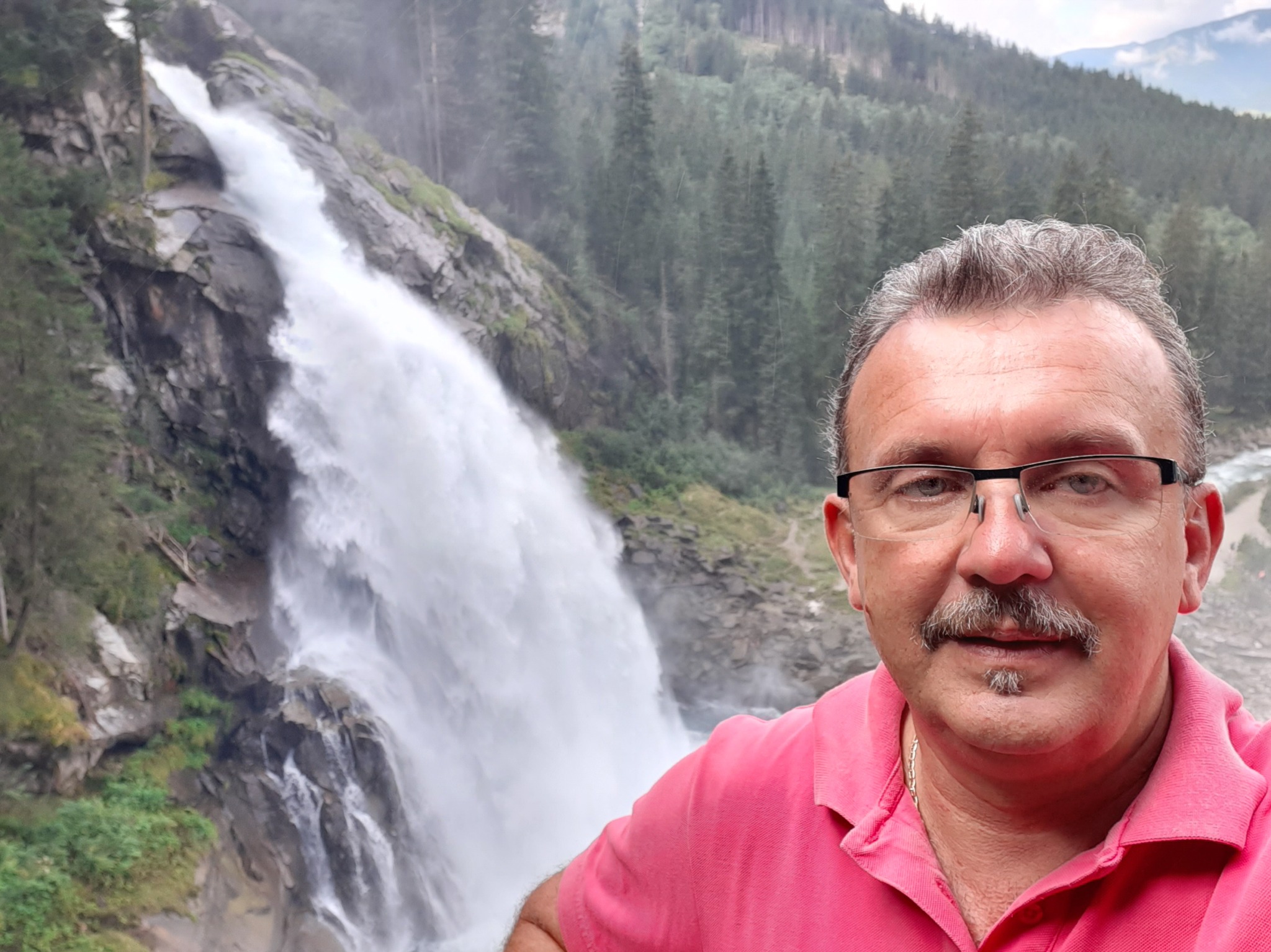Mężczyzna w różowej koszulce polo na tle wodospadu i gór porośniętych lasem.