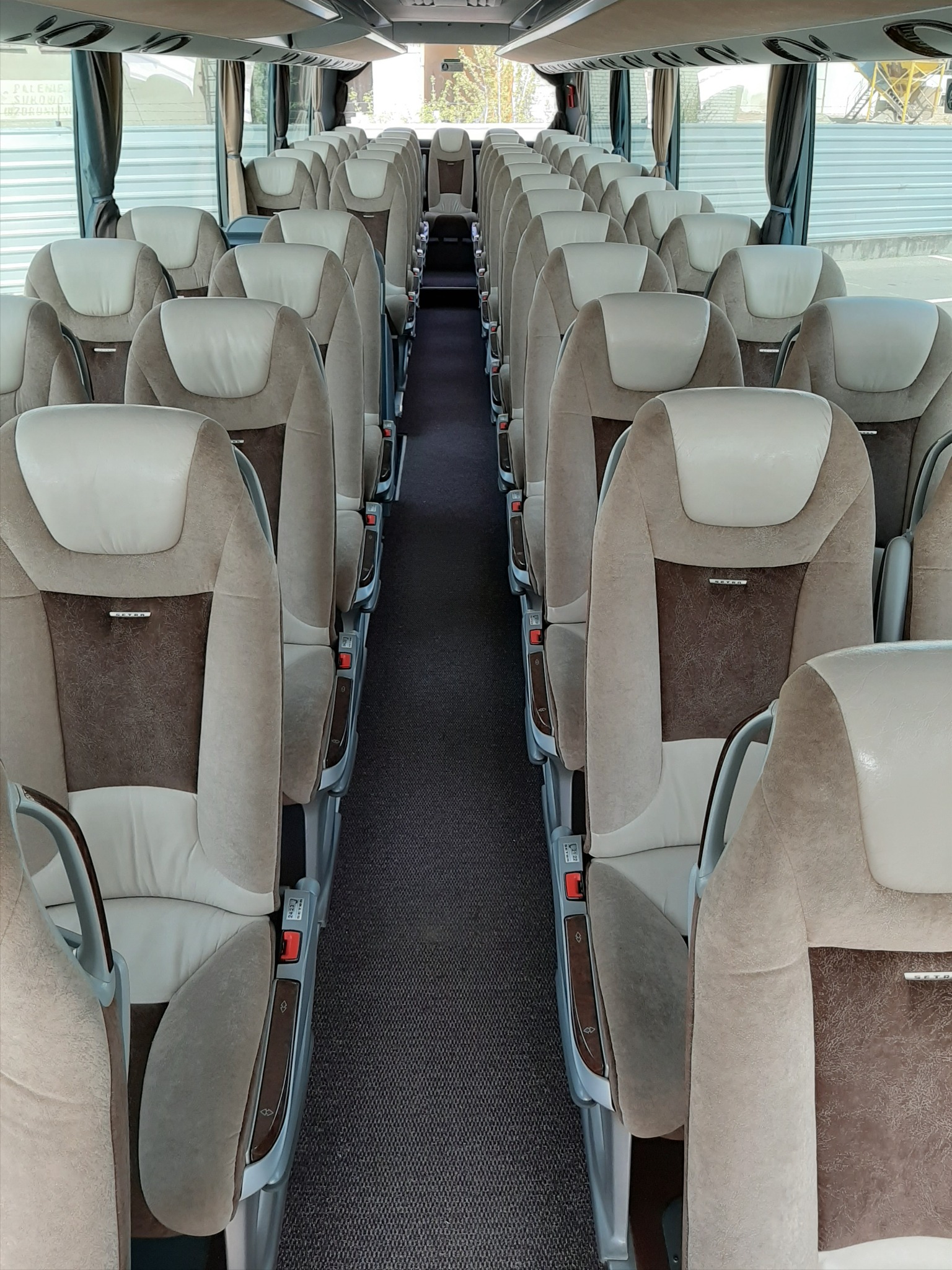 Wnętrze autobusu dalekobieżnego z rzędami beżowo-brązowych foteli z regulacją, widok z przodu kabiny pasażerskiej.