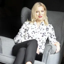 Kobieta z blond włosami siedzi w szarym fotelu, ubrana w białą koszulę w czarne kwiaty i czarne spodnie.