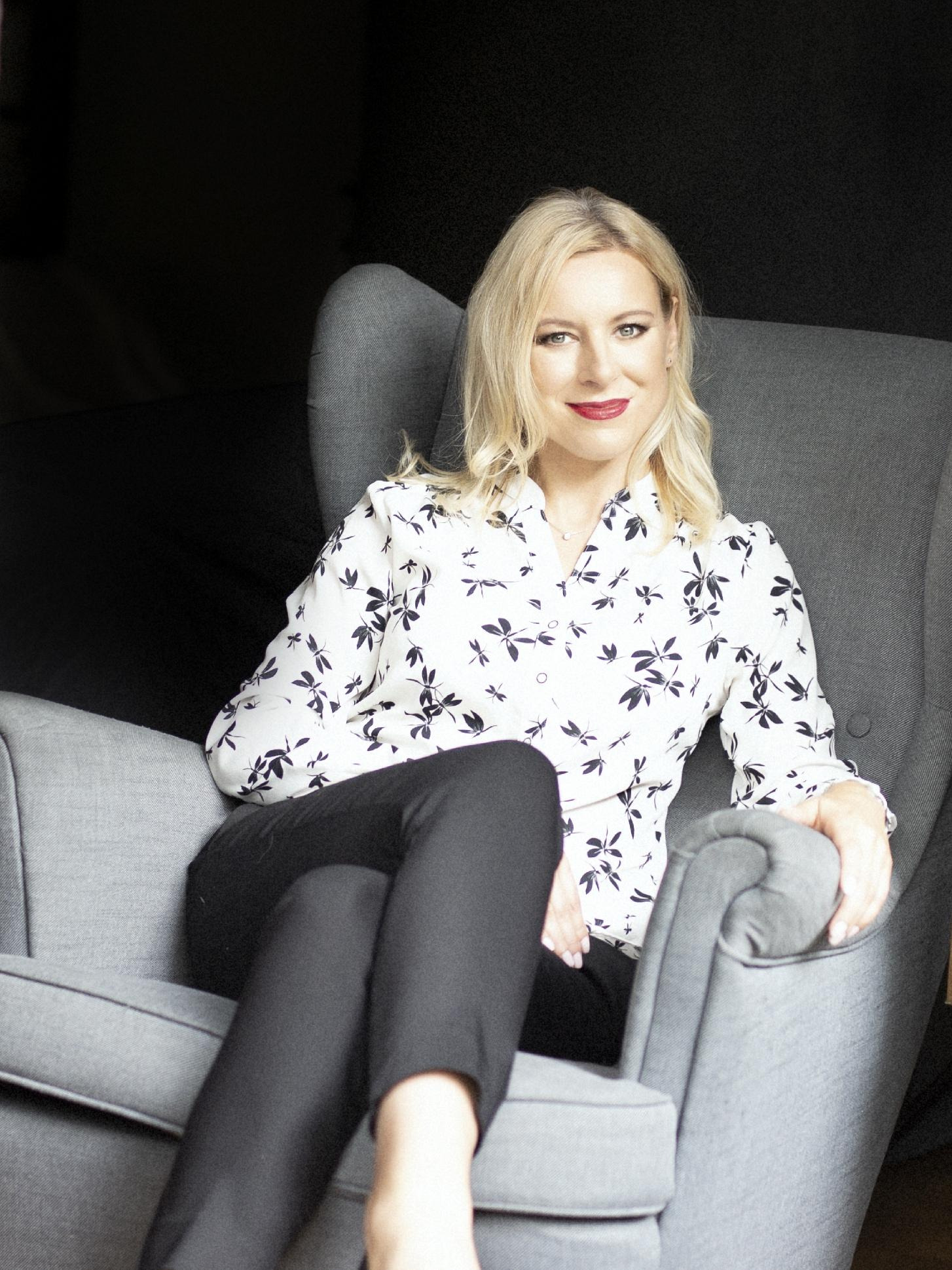 Kobieta z blond włosami siedzi w szarym fotelu, ubrana w białą koszulę w czarne kwiaty i czarne spodnie.