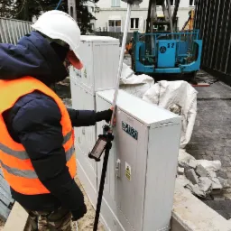 Pracownik w kasku i kamizelce mierzy położenie skrzynki elektrycznej za pomocą precyzyjnego odbiornika GNSS na tyczce, w tle widoczny dźwig.