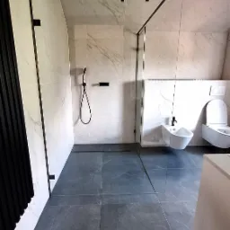 Nowoczesna łazienka z kabiną prysznicową typu walk-in, płytkami imitującymi marmur na ścianach, ciemnoszarą podłogą z odpływem liniowym, wiszącą toaletą i bidetem.