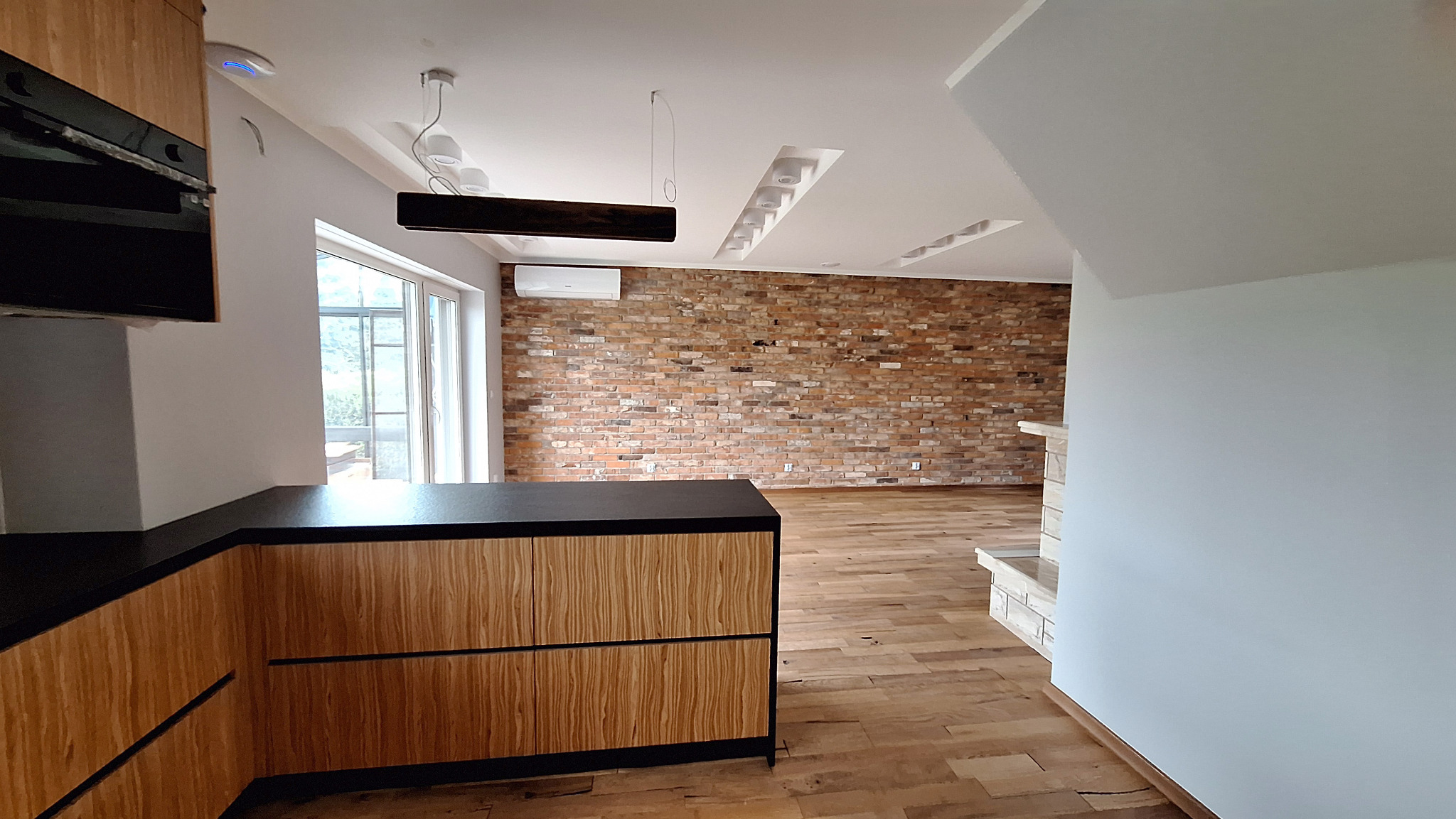 Nowoczesne wnętrze z drewnianą zabudową kuchenną, ceglaną ścianą i drewnianą podłogą. Widoczne minimalistyczne oświetlenie sufitowe i klimatyzator. W tle okno z widokiem na zieleń.
