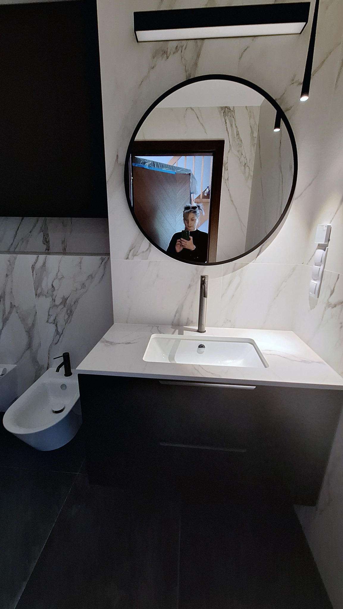 Nowoczesna łazienka z marmurowymi płytkami, okrągłym lustrem, czarną armaturą i bidetem, odbicie osoby robiącej zdjęcie w lustrze.