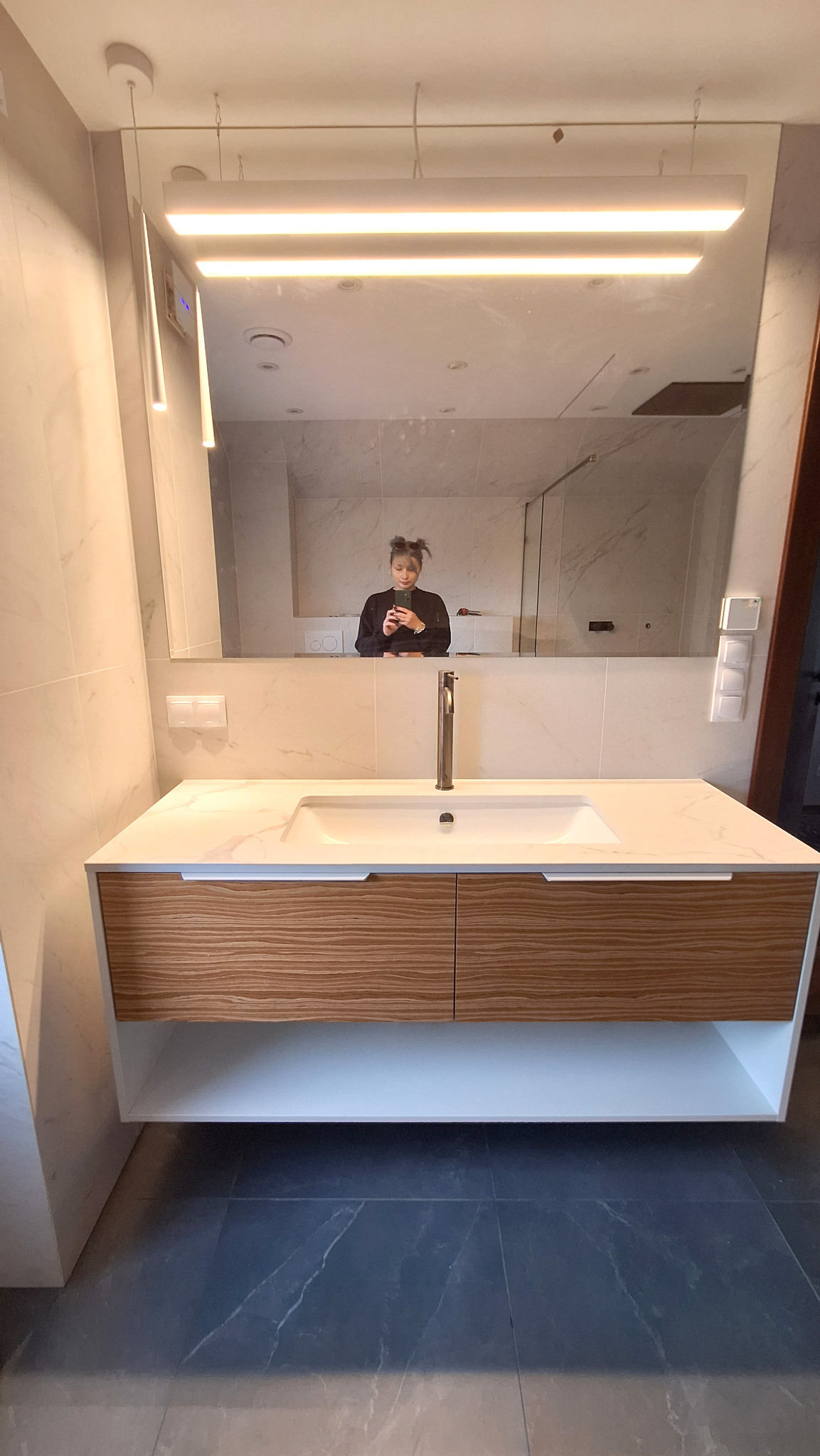 Nowoczesna łazienka z umywalką nablatową i szafką z drewnianymi frontami. W lustrze odbija się osoba robiąca zdjęcie telefonem. Widoczne oświetlenie LED nad lustrem i marmurowe płytki na ścianach.