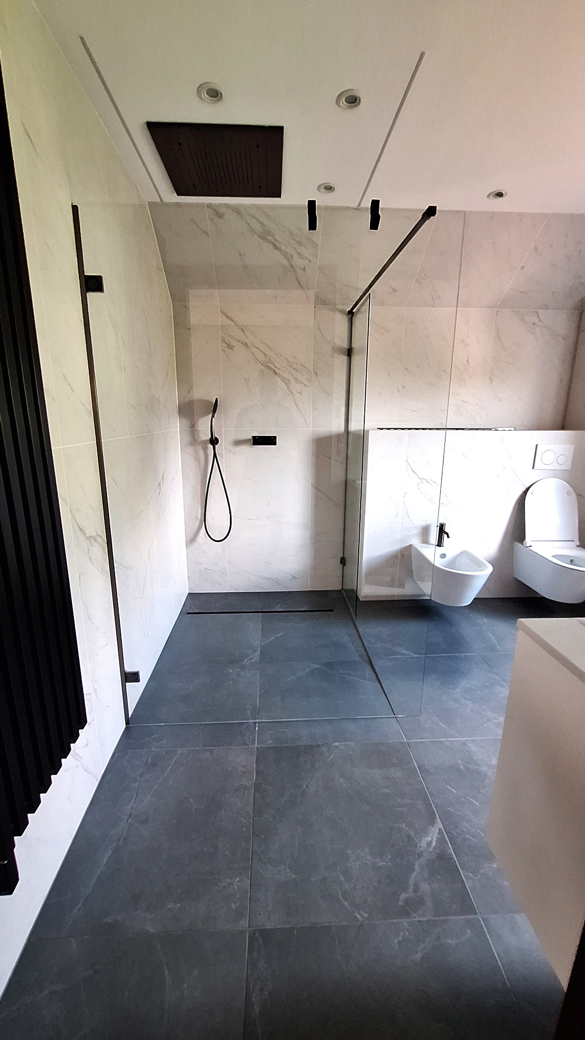 Nowoczesna łazienka z kabiną prysznicową typu walk-in, płytkami imitującymi marmur na ścianach, ciemnoszarą podłogą z odpływem liniowym, wiszącą toaletą i bidetem.