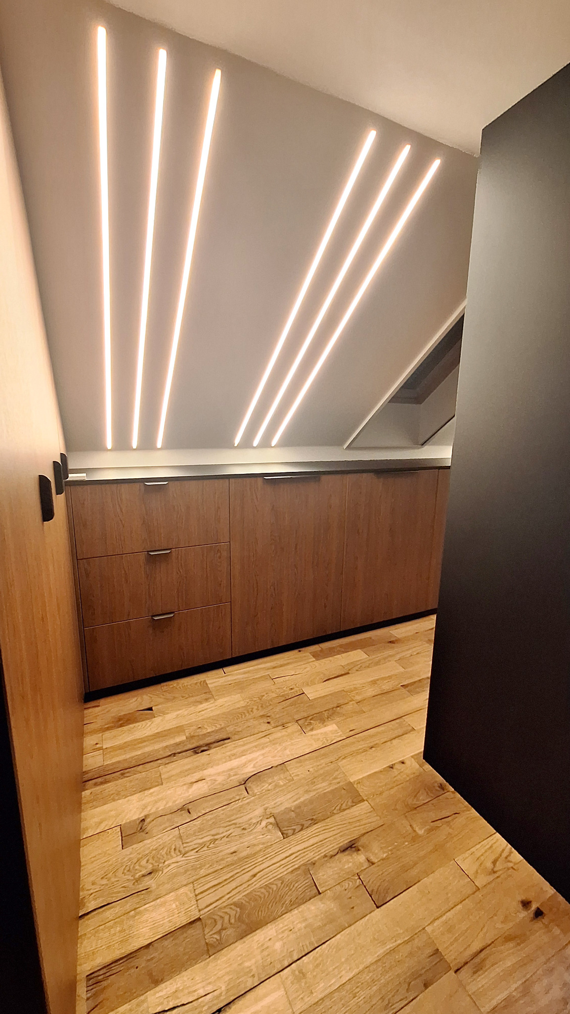 Nowoczesna aranżacja poddasza z zabudową kuchenną w odcieniach drewna, oświetleniem LED w suficie i drewnianą podłogą.