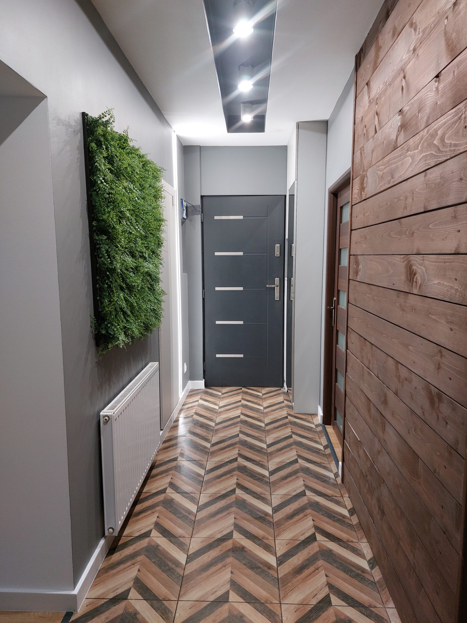 Wnętrze korytarza z drewnianą ścianą, podłogą w jodełkę, panelem z roślin na ścianie i szarą stolarką drzwiową.