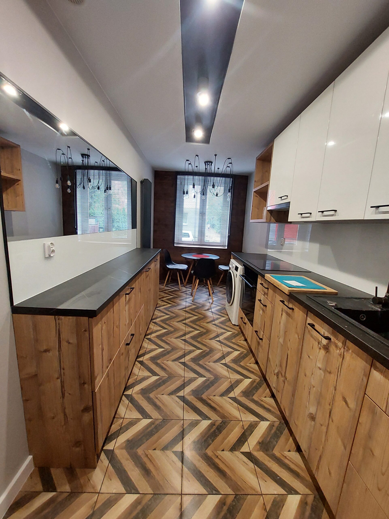 Wąska kuchnia w Bytomiu z drewnianymi szafkami dolnymi i białymi górnymi, czarnym blatem oraz wzorzystą podłogą z płytek. Na końcu pomieszczenia stół z krzesłami i nowoczesna lampa.