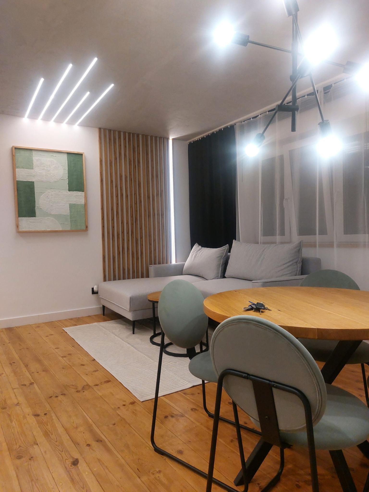 Jasne wnętrze mieszkania z drewnianą podłogą, szarą sofą i okrągłym drewnianym stołem z kluczami na blacie, oświetlone nowoczesną lampą sufitową i ledowymi paskami, z obrazem w zielonej ramie...