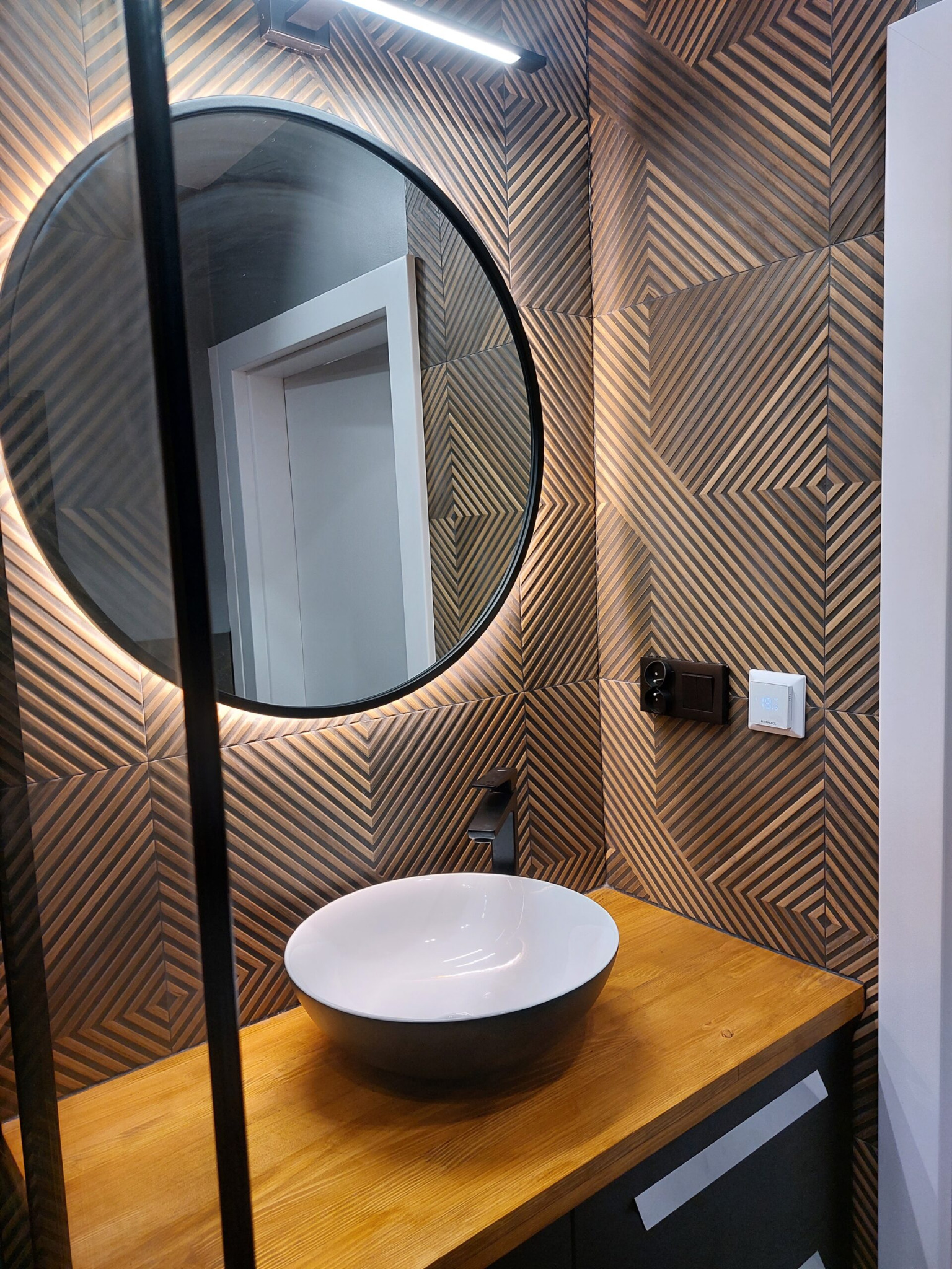 Nowoczesna łazienka z okrągłym lustrem podświetlanym LED, umywalką nablatową na drewnianym blacie i dekoracyjną ścianą z drewnianych paneli z geometrycznym wzorem.