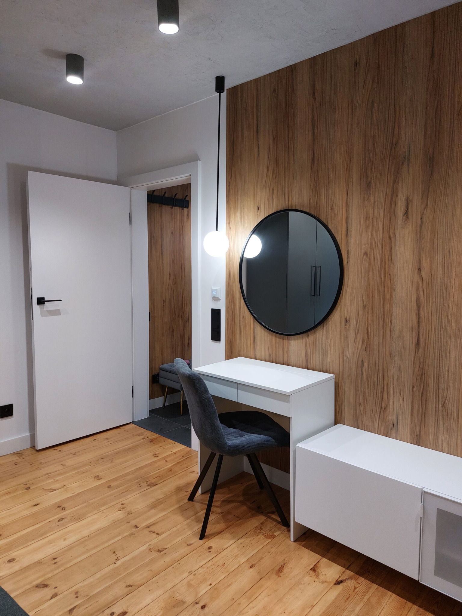 Nowoczesna toaletka z okrągłym lustrem na tle ściany z drewnopodobną okładziną, szara tapicerowana krzesło i biała szafka RTV na drewnianej podłodze, oświetlenie punktowe w suficie.
