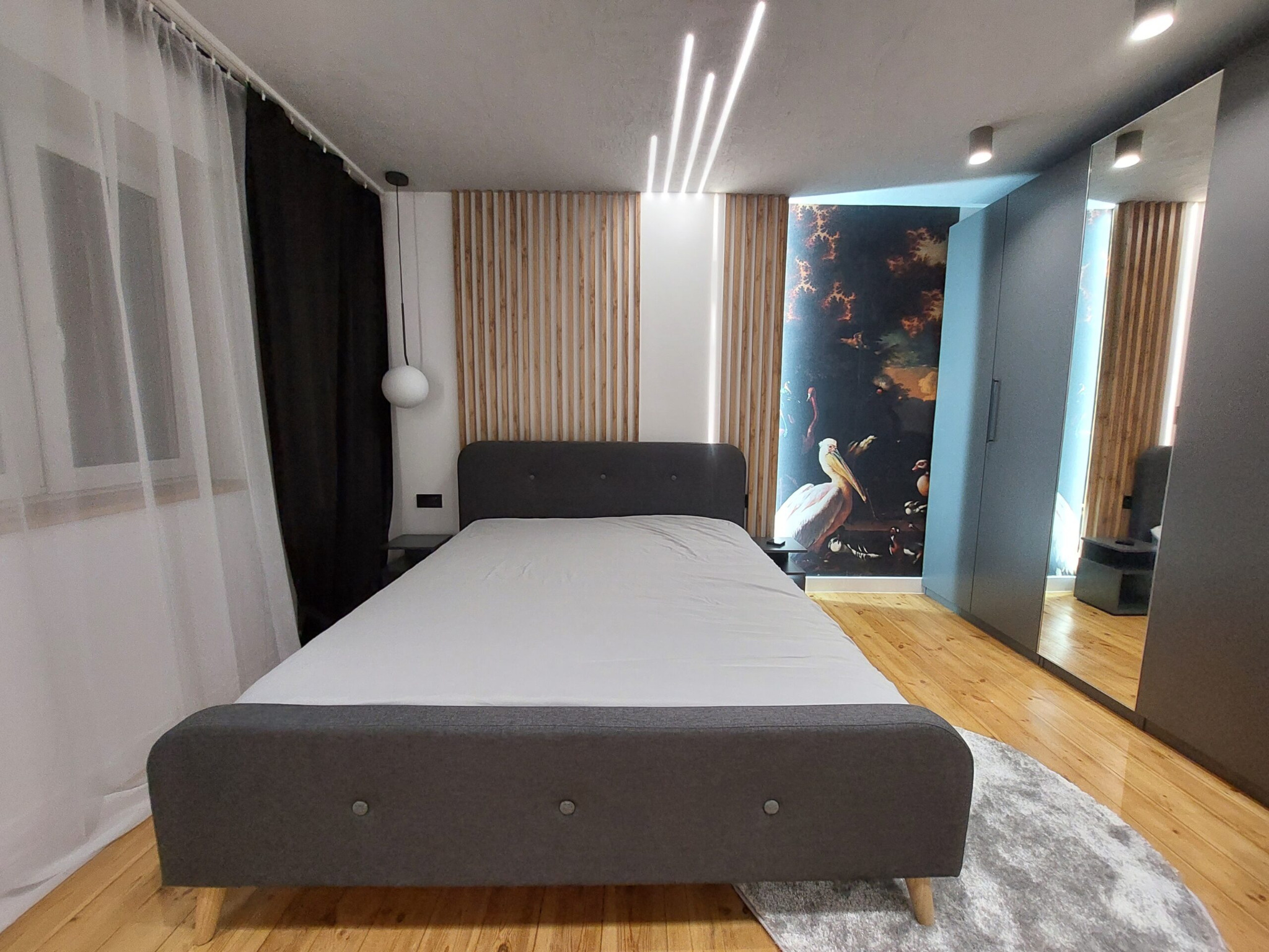 Sypialnia z szarą tapicerowaną ramą łóżka, drewnianymi lamelami na ścianie, fototapetą z motywem barokowym i wysoką szafą z lustrem.