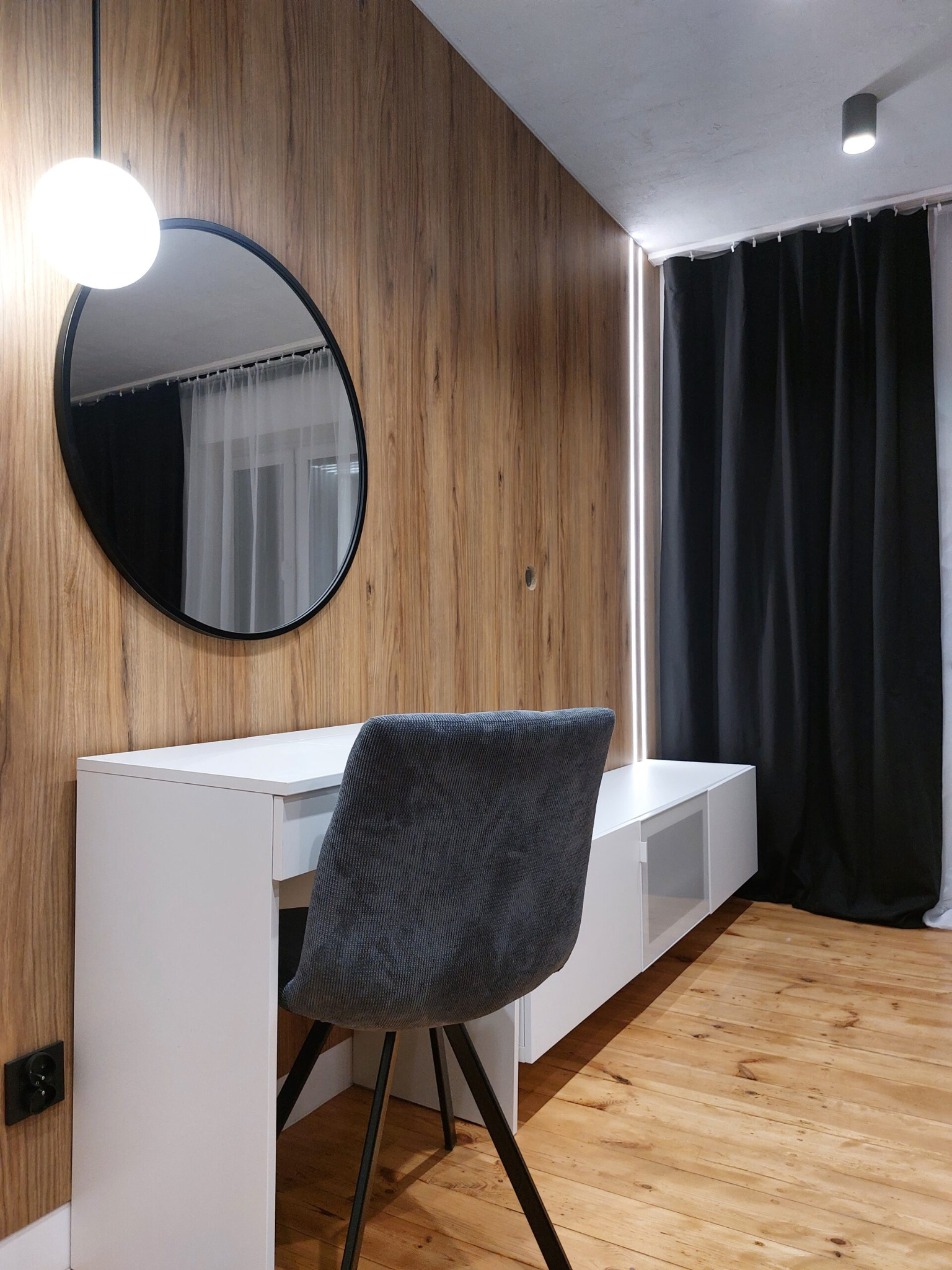 Nowoczesna aranżacja wnętrza z biurkiem, okrągłym lustrem i drewnianą ścianą, uzupełniona o czarne zasłony i podświetlenie LED.