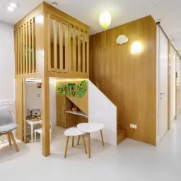 Jasne wnętrze kliniki dentystycznej w Warszawie z drewnianą konstrukcją do zabaw dla dzieci, w tle widoczny korytarz z gabinetami.