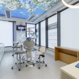 Jasne wnętrze gabinetu z nowoczesnym fotelem dentystycznym, szafkami z białymi frontami i sufitem z motywem kwitnących gałęzi, oświetlenie LED w kształcie okręgu.