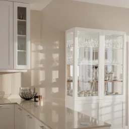 Jasna, elegancka kuchnia z białą zabudową, przeszkloną witryną wypełnioną szkłem i porcelaną, oraz blatem z delikatnym połyskiem, oświetlona naturalnym światłem padającym przez okno.