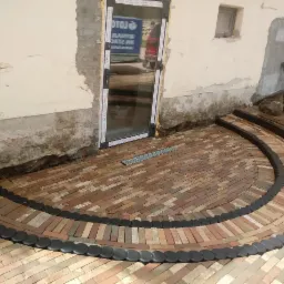 Półkolisty taras z ceglanej kostki, obrzeża z ciemnych, okrągłych elementów, przy ścianie budynku z widocznymi śladami prac budowlanych i nowymi oknami.