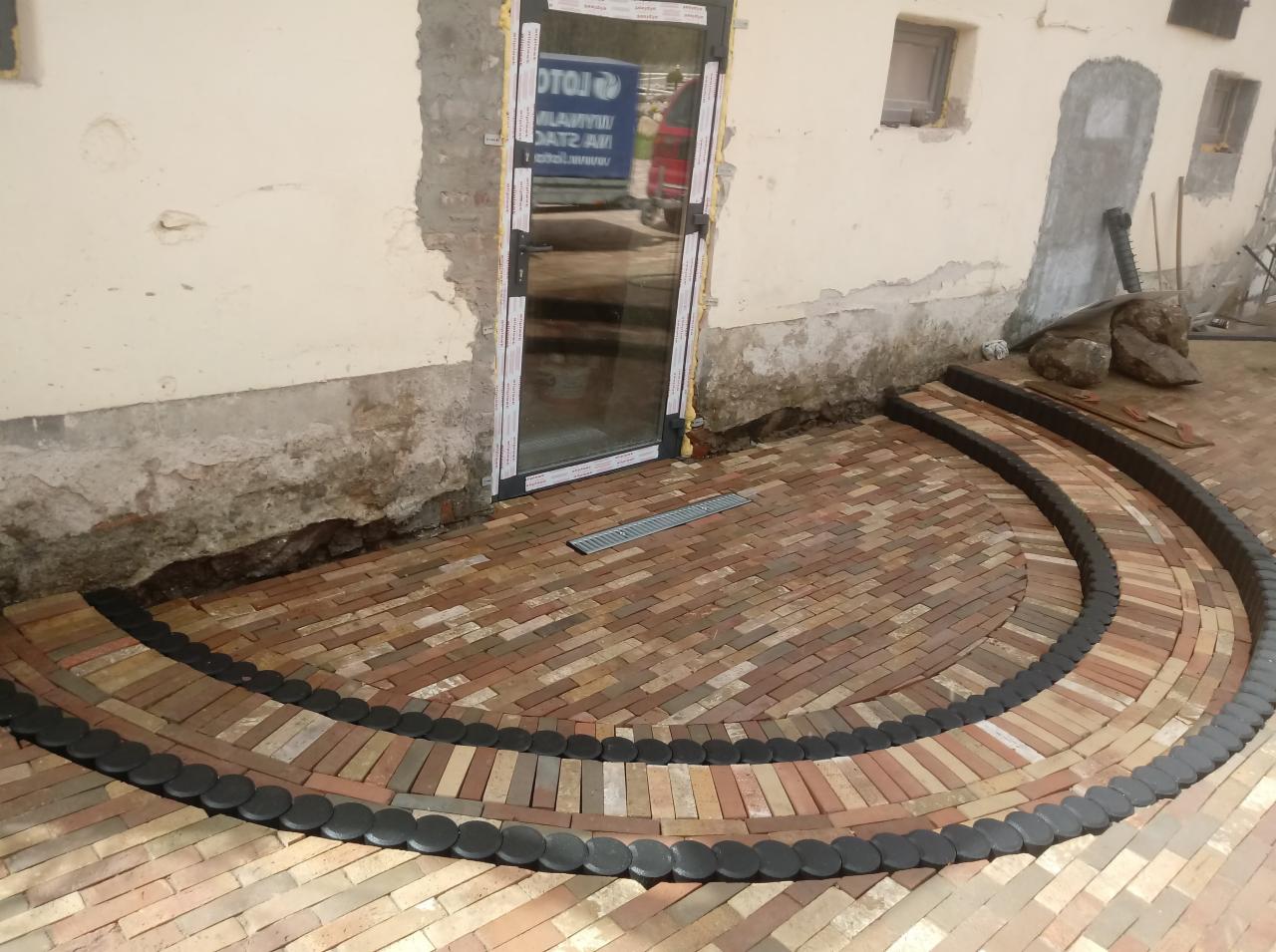 Półkolisty taras z ceglanej kostki, obrzeża z ciemnych, okrągłych elementów, przy ścianie budynku z widocznymi śladami prac budowlanych i nowymi oknami.