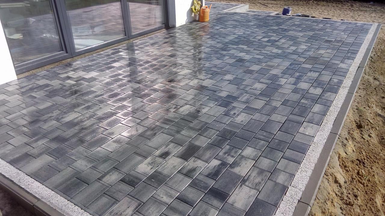 Ułożona, szara kostka brukowa w jodełkę na tarasie domu, z widocznym obrzeżem i elementami wykończeniowymi, częściowo mokra po deszczu, w tle okno tarasowe i doniczka.