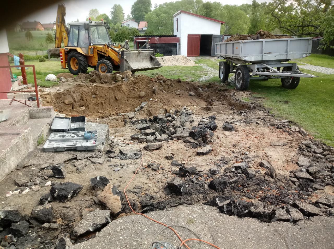 Żółta koparko-ładowarka i przyczepa z ziemią w trakcie prac ziemnych na posesji, widoczne rozbite fragmenty starej nawierzchni i wykop pod przyszłą instalację.