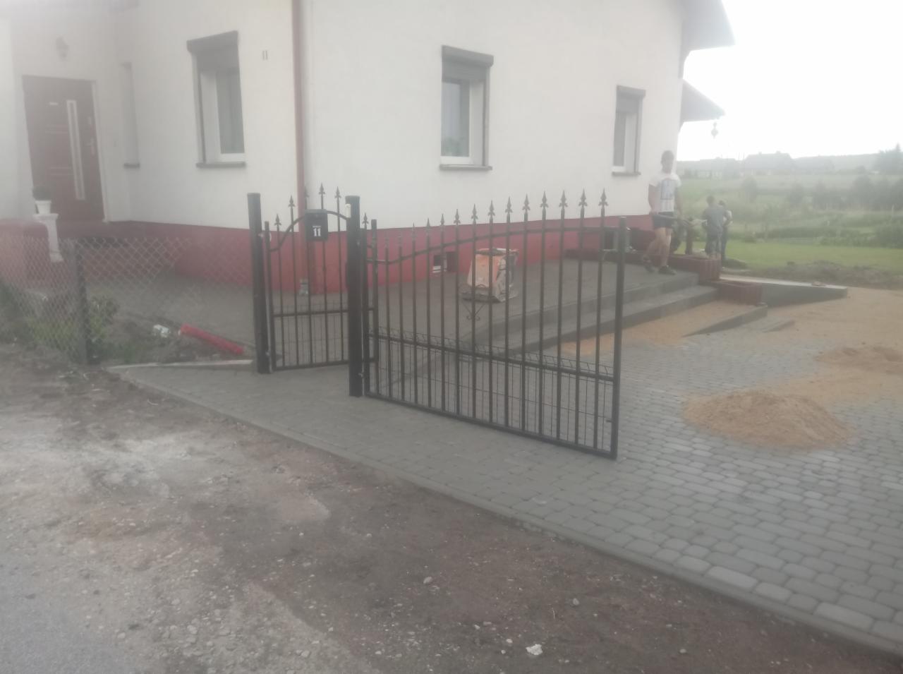 Nowo ułożona kostka brukowa przed domem z zamontowaną, czarną, metalową bramą z dekoracyjnymi grotami na górze, przy której leży rozsypany piasek do spoinowania.
