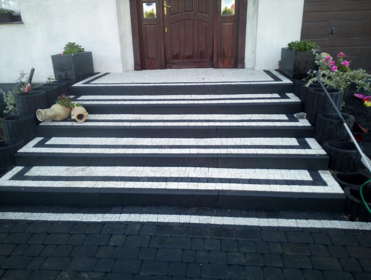 Schody wejściowe wyłożone czarno-białą kostką, prowadzące do drzwi wejściowych, z dekoracyjnymi donicami po bokach i fragmentem podjazdu z ciemnej kostki brukowej.