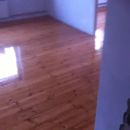 Błyszcząca, odnowiona drewniana podłoga w pokoju, widoczne ślady po cyklinowaniu i świeżo nałożony lakier, odbijający światło z okna.