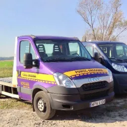 Dwa samochody: fioletowa laweta Iveco z żółtym pasem 'Pomoc Drogowa' i granatowy van Fiat Ducato na polnej drodze w słoneczny dzień.