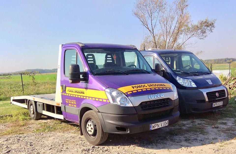 Dwa samochody: fioletowa laweta Iveco z żółtym pasem 'Pomoc Drogowa' i granatowy van Fiat Ducato na polnej drodze w słoneczny dzień.