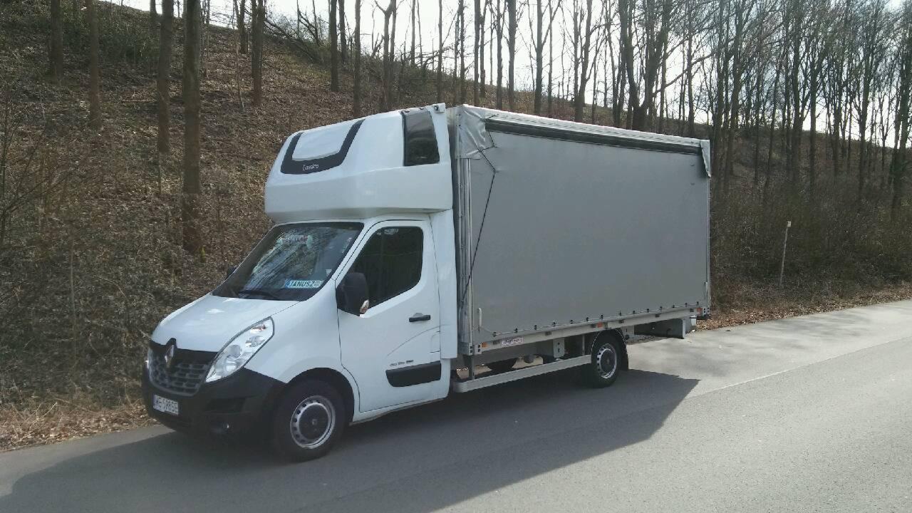 Biały Renault Master z zabudową plandekową typu firanka, zaparkowany na poboczu drogi w pobliżu lasu, widoczna tablica rejestracyjna.