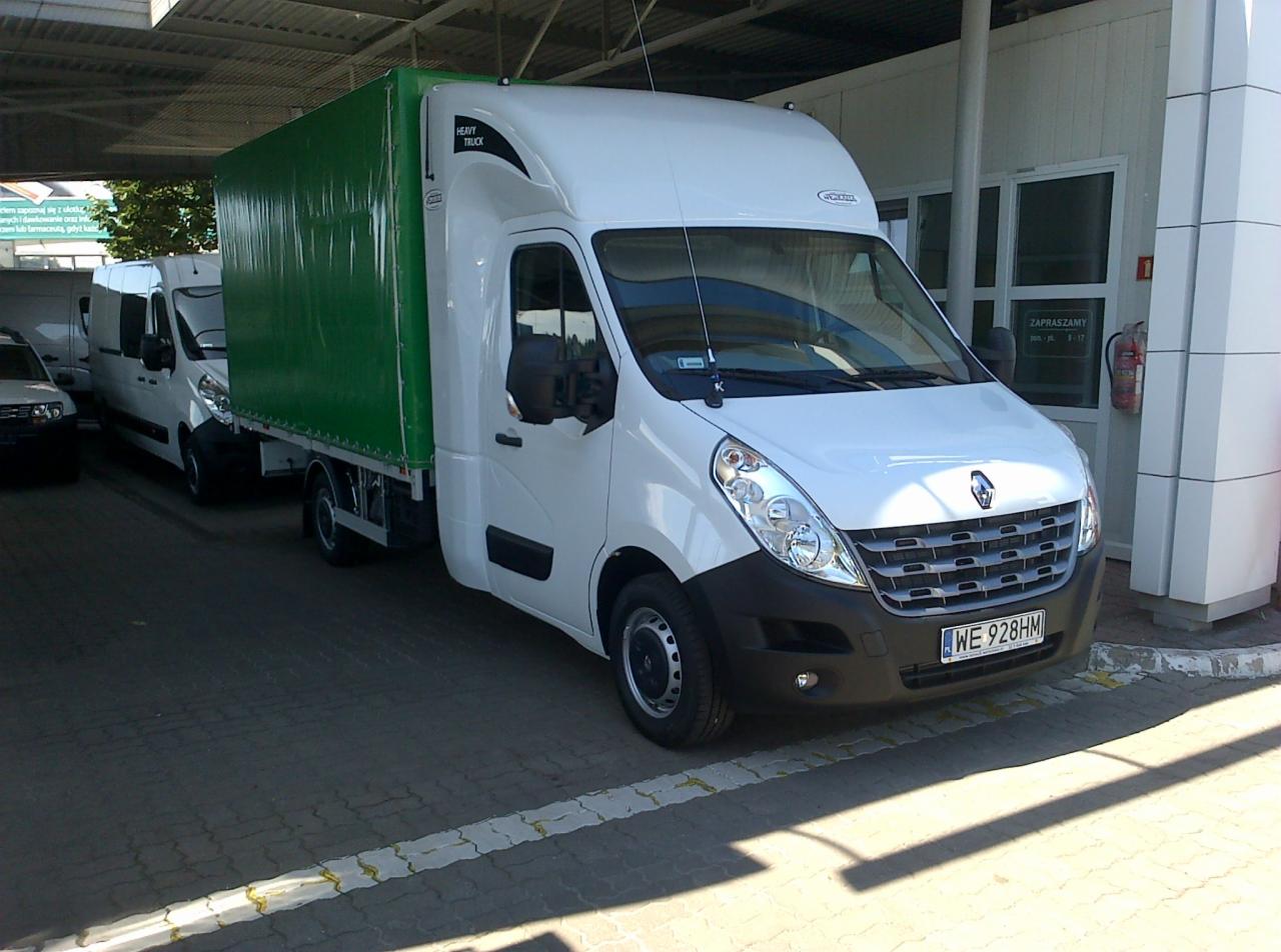 Biały bus dostawczy Renault Master z zieloną plandeką, zaparkowany na kostce brukowej przed budynkiem z napisem 'Zapraszamy', widziany z przodu pod lekkim kątem.