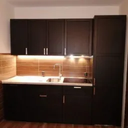 Ciemna zabudowa kuchenna z górnymi szafkami, blatem z umywalką i płytą grzewczą oraz podświetleniem LED podszafkowym na tle płytek imitujących drewno.